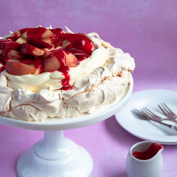 A pavlova topped with peaches and raspberry sauce on a white cake stand, a small jug of raspberry sauce and a white plate and cake forks on a pink background