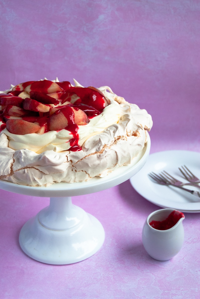 A pavlova topped with peaches and raspberry sauce on a white cake stand, a small jug of raspberry sauce and a white plate and cake forks on a pink background