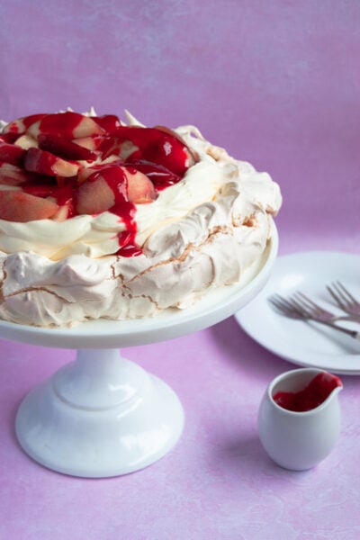 A pavlova topped with whipped cream, fresh sliced peaches and raspberry sauce on a white cake stand, a small white jug of raspberry sauce and white plates and cake forks on a pink background