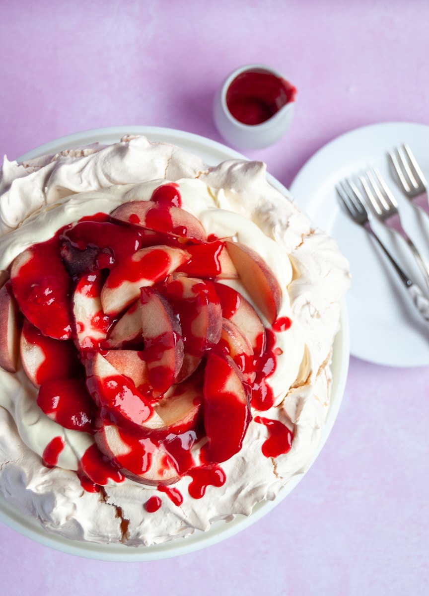 A flat lay photo of a peach melba pavlova topped with raspberry sauce on a white cake stand, a small jug of raspberry sauce and a white plate and cake forks