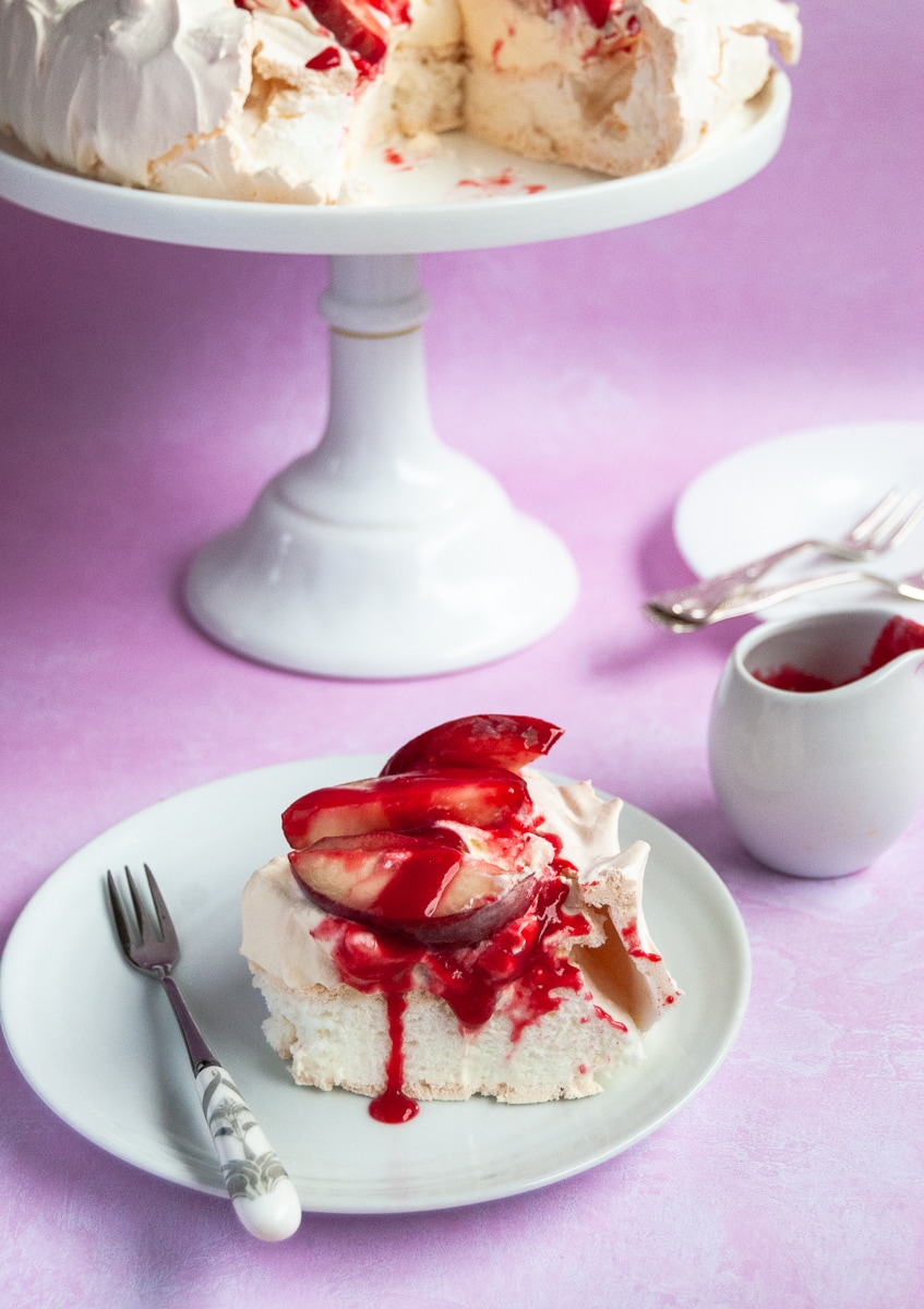 A slice of peach melba pavlova topped with peaches and raspberry sauce, a cake stand with a pavlova, a small jug of raspberry sauce and a white plate and cake forks on a pink background