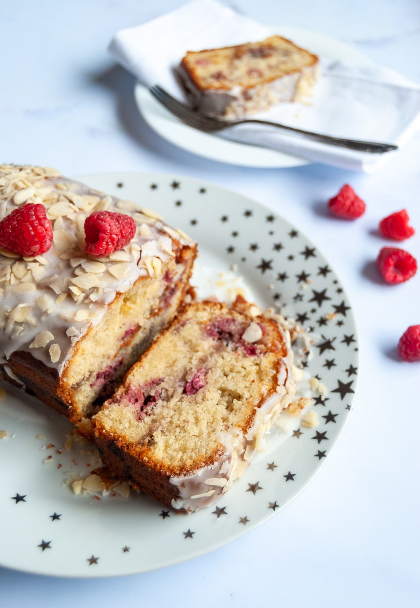 A raspberry Bakewell cake topped with white icing, flaked almonds and fresh raspberries on a white and gold star plate and a slice of the cake on a small white plate with a cake fork