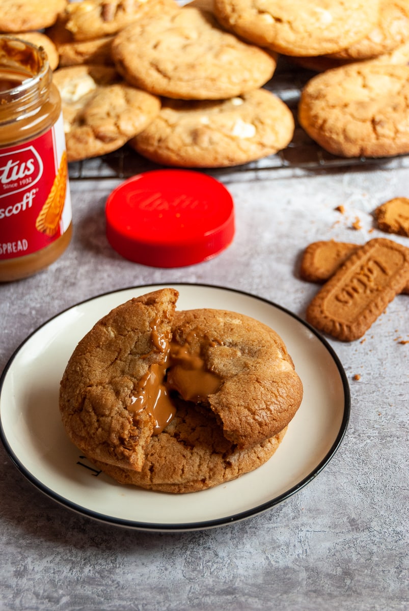 A biscoff cookie on a white and black plate broken in the middle to reveal a molten Biscoff centre. More cookies and a jar of Biscoff in the background