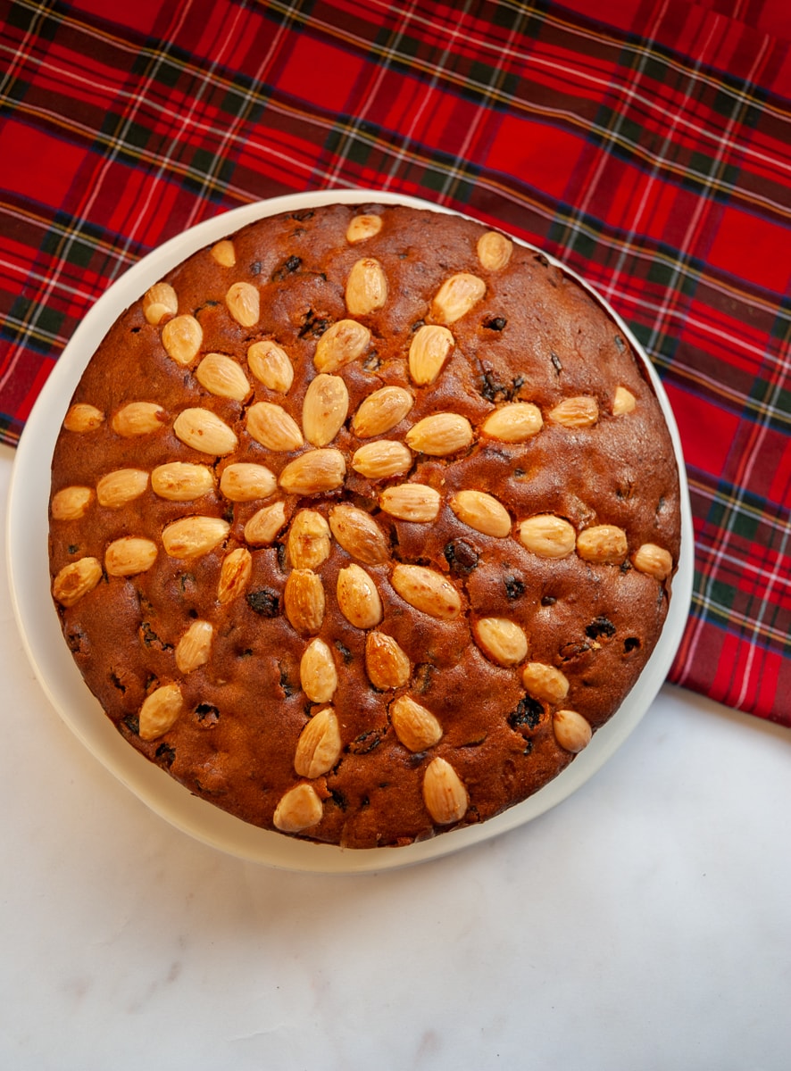 A fruit Dundee cake studded with whole almonds on a white plate and a red tartan tablecloth