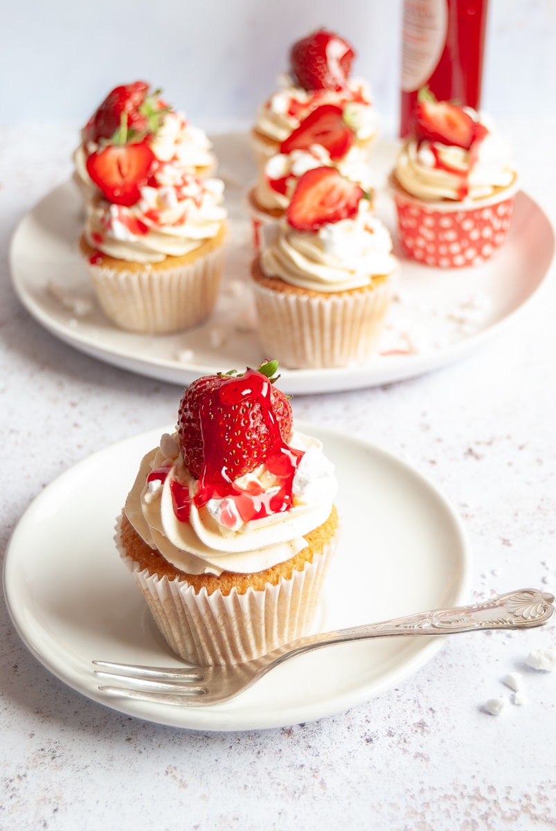 An Eton Mess Cupcake topped with a strawberry on a white plate with a cake fork.