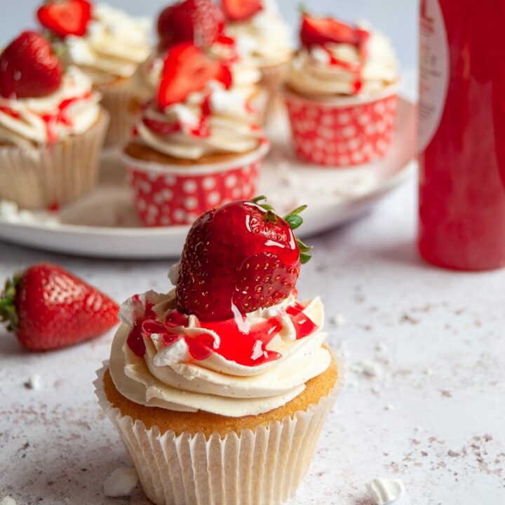 A vanilla cupcake with vanilla buttercream, crushed meringue and a strawberry. A plate of cupcakes can be seen on a white plate in the background and a bottle of strawberry sauce