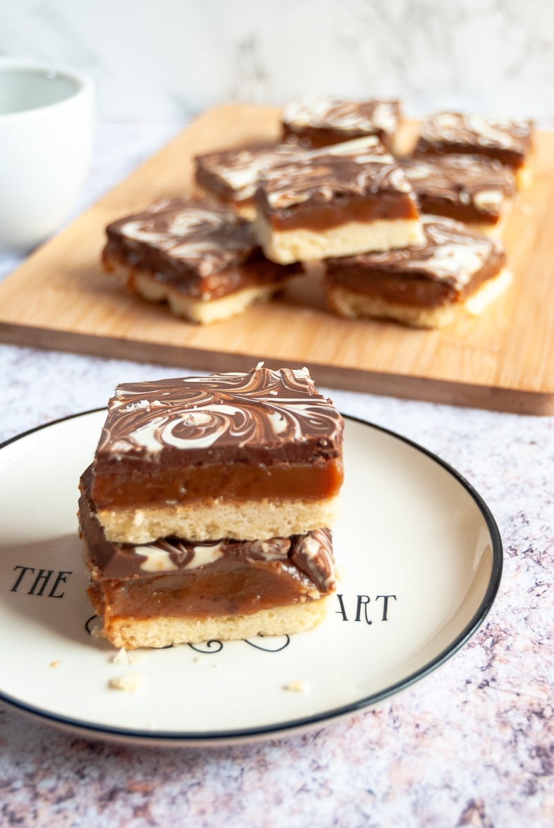 two pieces of millionaires shortbread stacked on top of each other on a white and black plate. A wooden board with more caramel shortbread in the background.