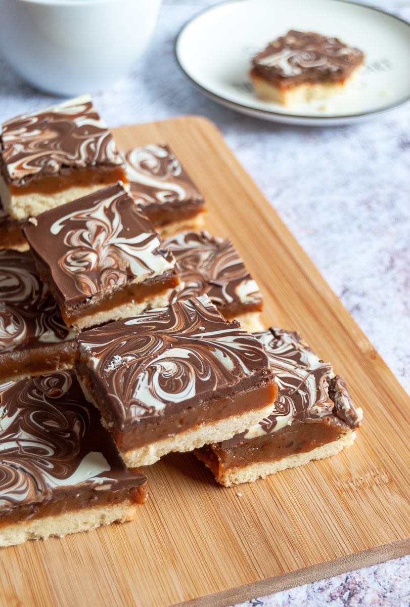 squares of chocolate topped caramel shortbread on a wooden board, a white coffee cup and a plate with a small piece of shortbread