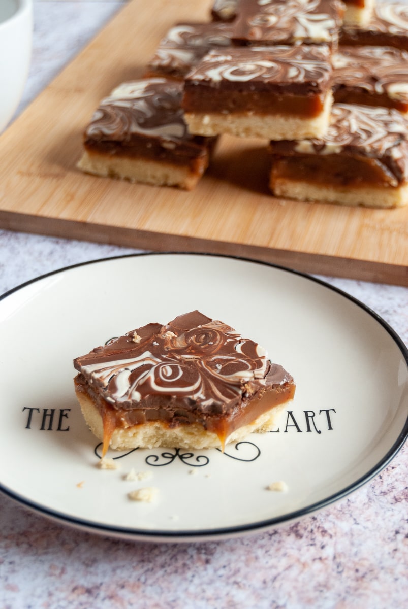 A piece of millionaires shortbread on a white and black plate, with more pieces of the shortbread on a wooden board