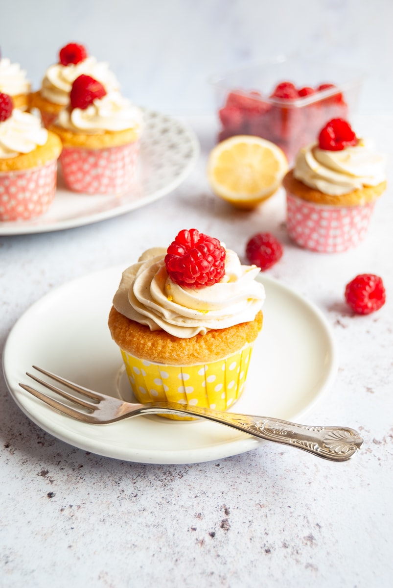 A lemon cupcake topped with lemon buttercream and a fresh raspberry on a white plate with a cake fork. More cupcakes can be seen in the background with a punnet of raspberries and half a lemon.