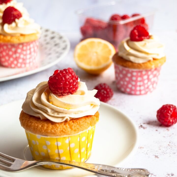A raspberry and lemon cupcake on a white plate with a cake fork on a grey and white background. More cupcakes can be partially seen in the background.