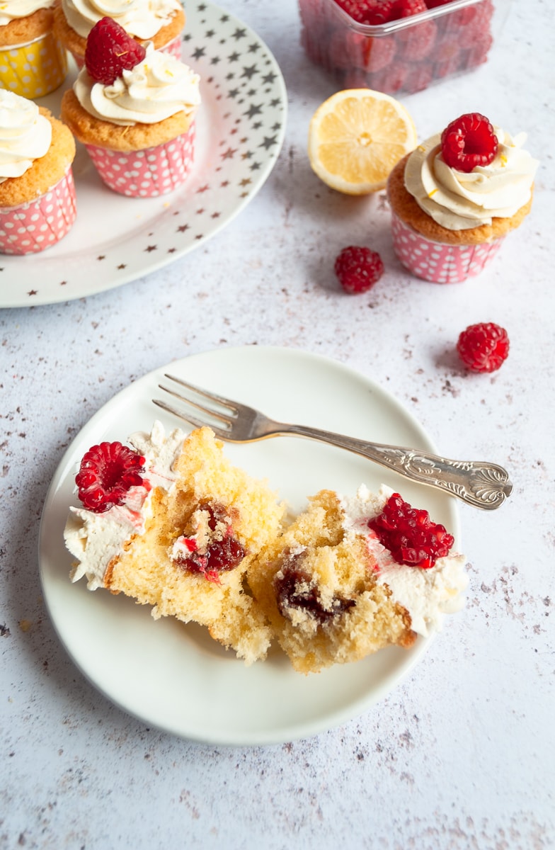 A lemon cupcake on a white plate with a cake fork, cut in half to reveal the jam middle. more cupcakes can be seen in the top left corner 