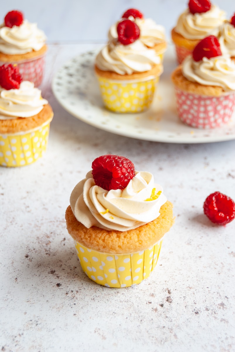 A raspberry and lemon cupcake with a lemon buttercream swirl and topped with a fresh raspberry on a grey and white background 