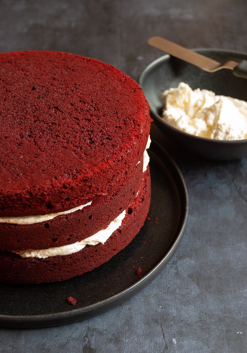 A 3 layer red velvet cake on a black plate and a small black bowl of white chocolate frosting