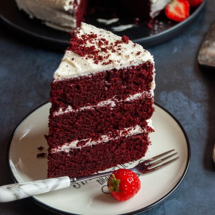 A slice of red velvet cake on a white and black plate with a cake fork and a fresh strawberry