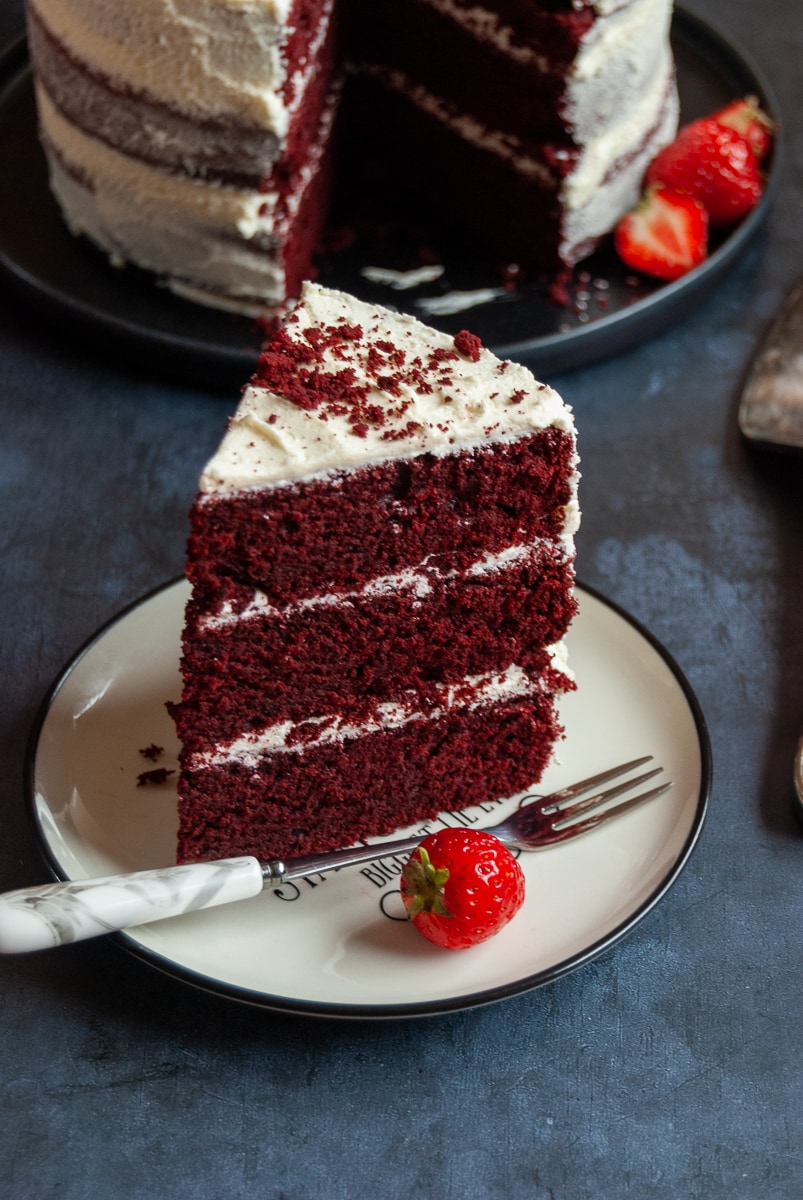 A slice of red velvet cake on a white and black plate with a cake fork and a fresh strawberry