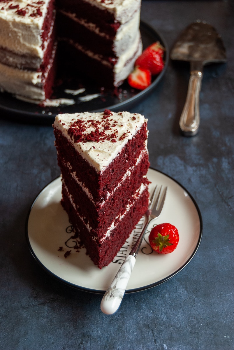 a slice of red velvet cake on a white and black plate with a cake fork and a fresh strawberry. 