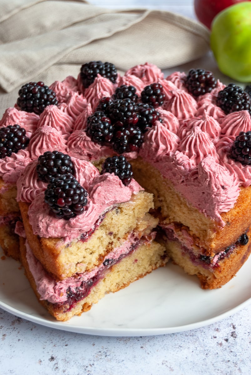 a close up photo of an apple cake with mixed berry jam, blackberry buttercream and topped with fresh blackberries on a white plate.