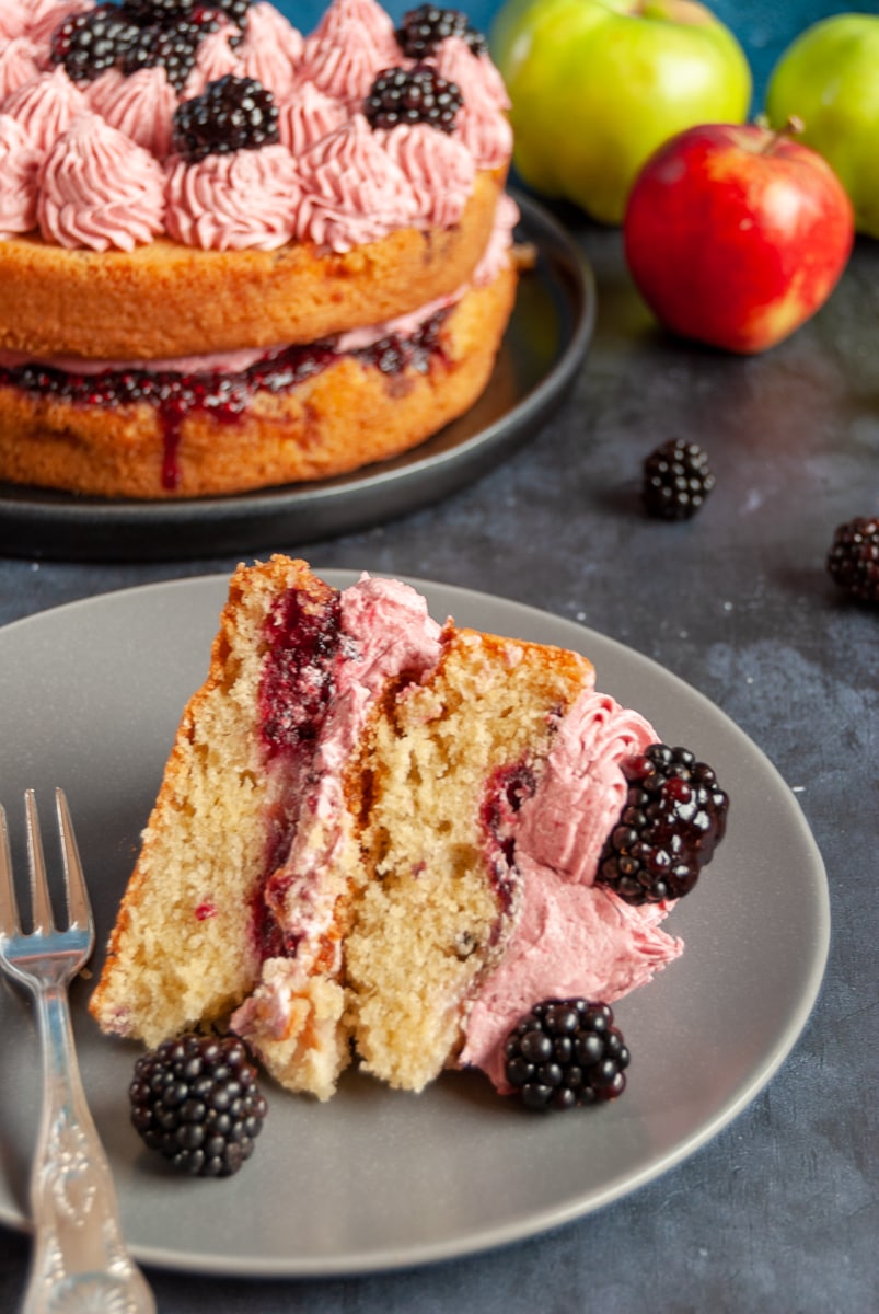 A slice of apple and blackberry cake on a grey plate and a large cake with blackberry buttercream and fresh blackberries in the background