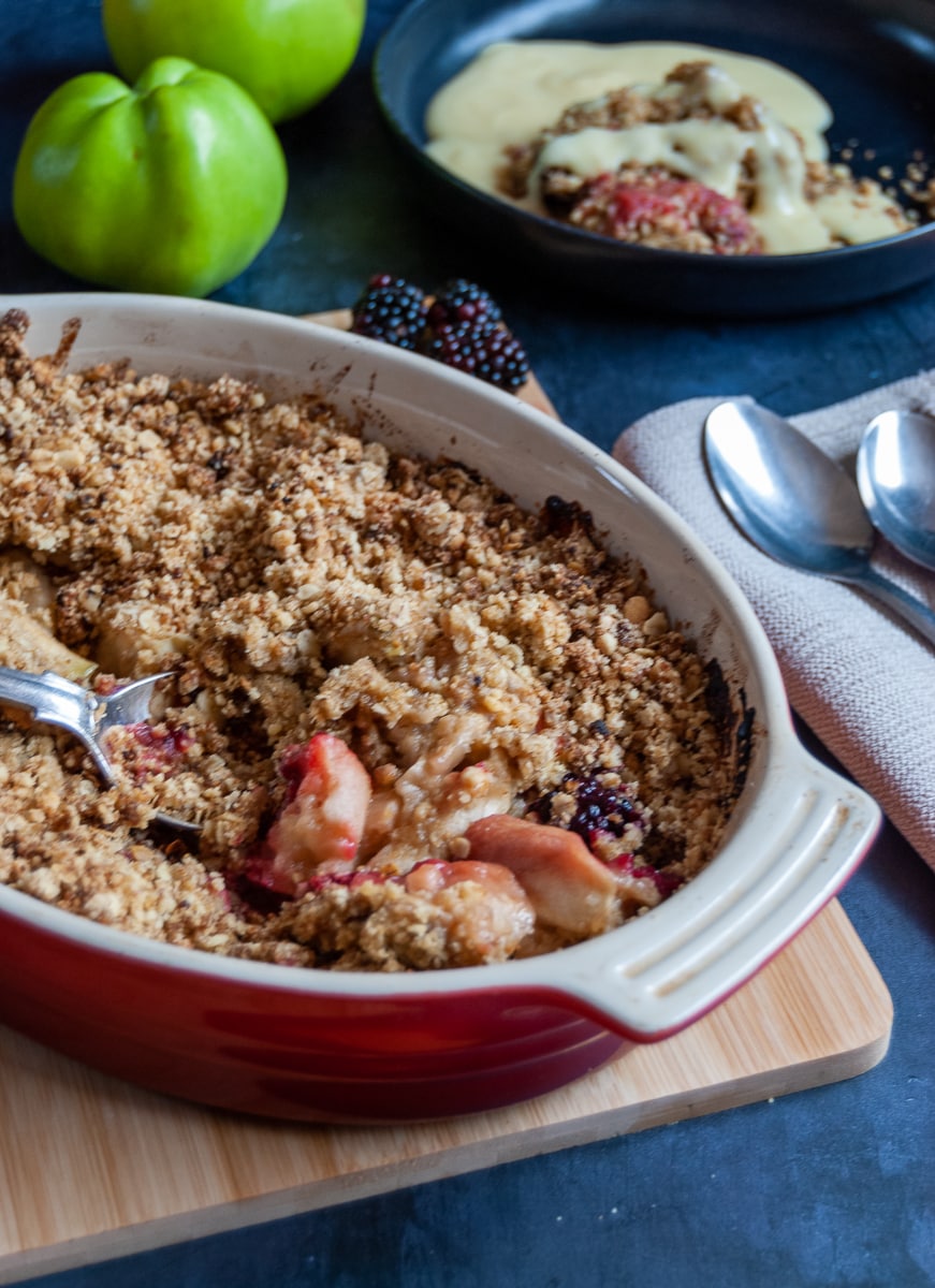 A close up picture of an apple and blackberry Crumble in a red oval dish on a wooden board, napkins and spoons and a black bowl of the crumble with custard in the background.