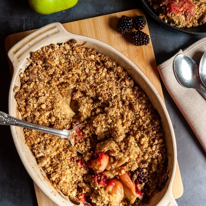 a dish of apple and blackberry crumble on a wooden board, a small bowl of the crumble and green apples and blackberries beside the dish.
