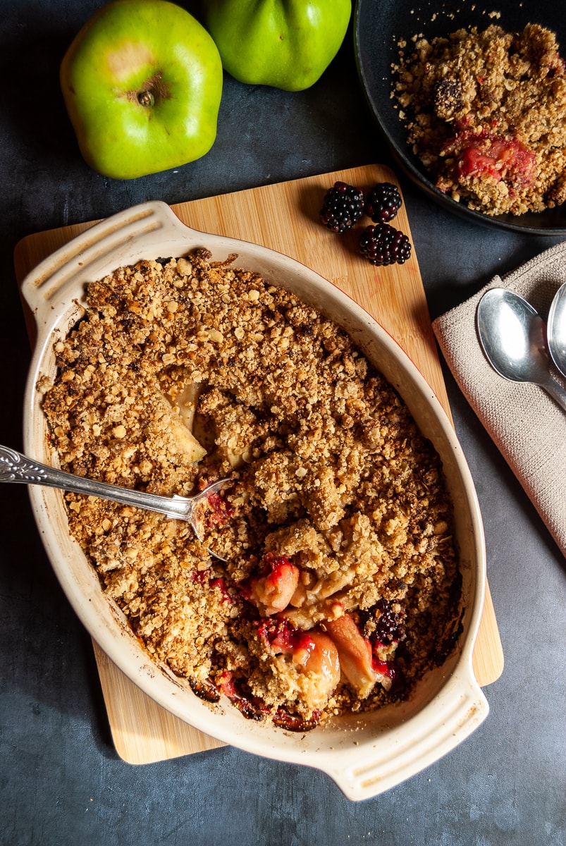 a dish of apple and blackberry crumble on a wooden board, a small bowl of the crumble and green apples and blackberries beside the dish.