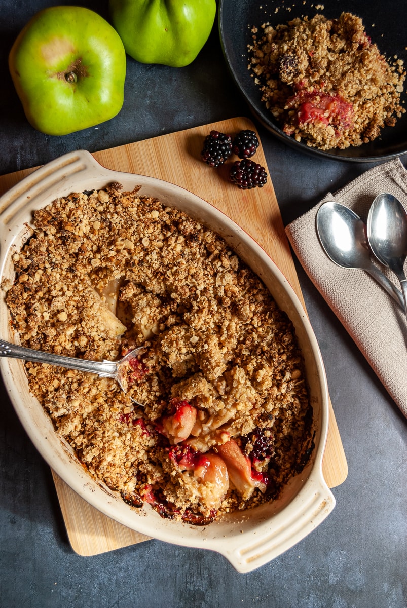 A homemade apple and blackberry Crumble in an oval dish on a wooden chopping board, two spoons and a napkin and a small bowl of the Crumble 