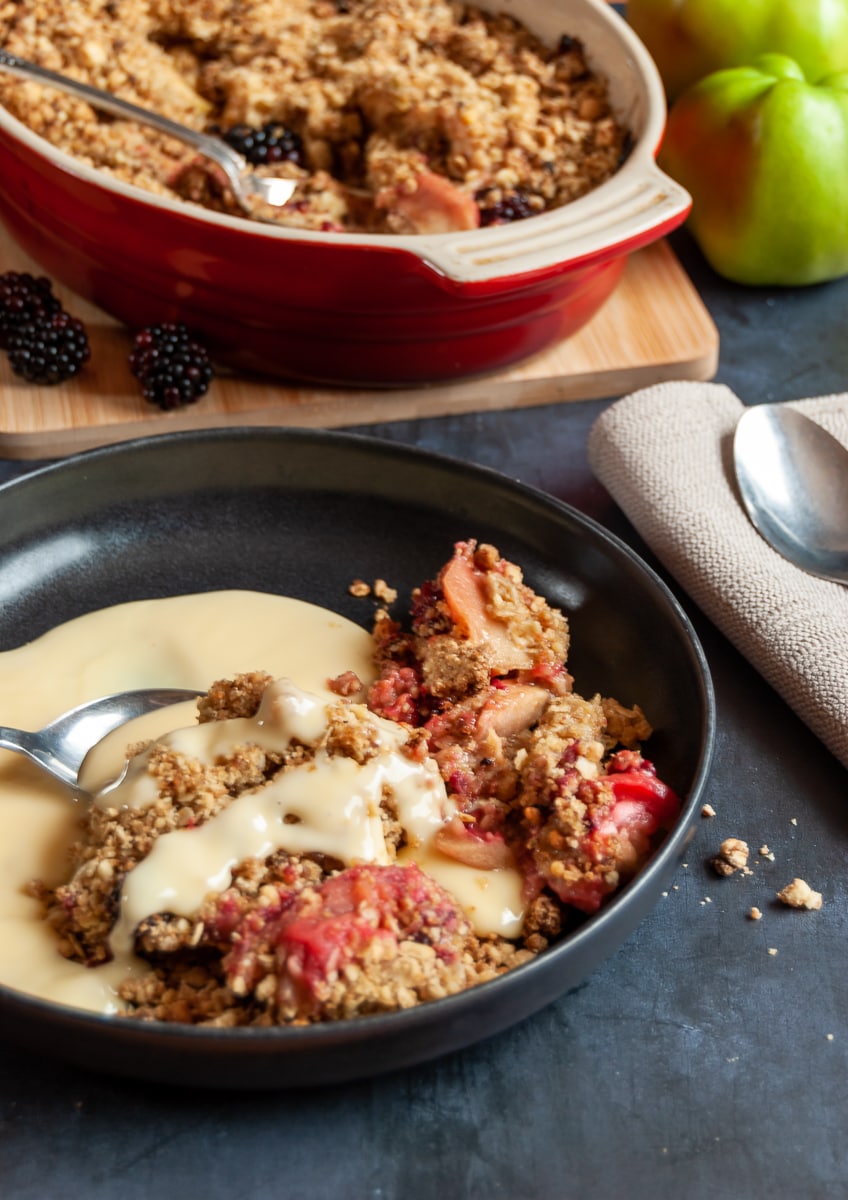 A black bowl of apple and blackberry crumble with custard and a spoon and a large dish of crumble on a wooden board on the background