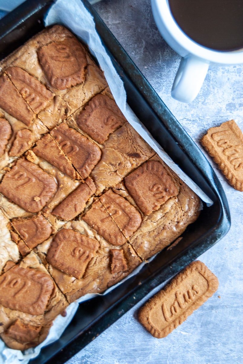 a flat lay photo of a tray of biscoff blondies, a white cup of coffee and Lotus biscuits