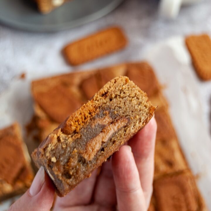 a hand holding a Biscoff stuffed blondie and more blondies in the background with a white cup of coffee