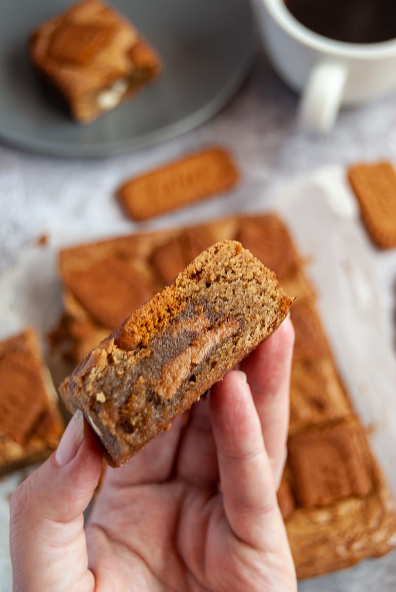 a hand holding a Biscoff stuffed blondie and more blondies in the background with a white cup of coffee