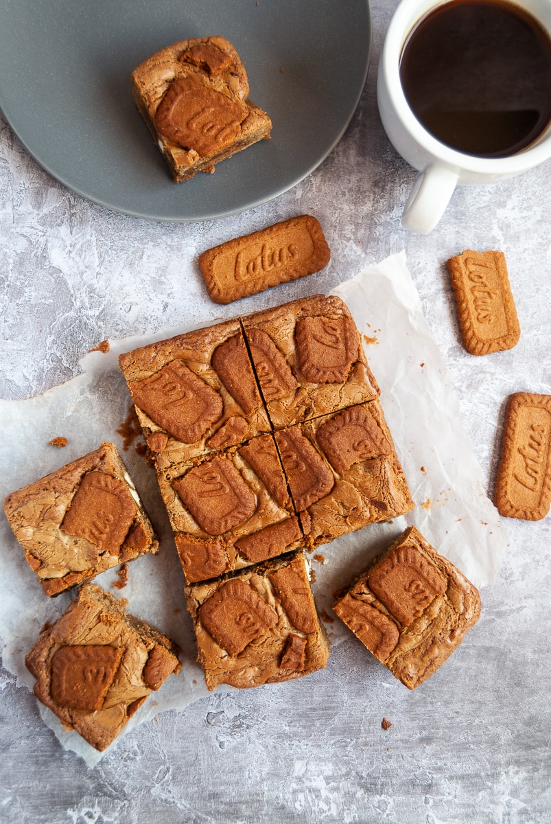 Biscoff blondies on a piece of baking parchment, a white cup of black coffee and a grey plate with a biscoff blondie