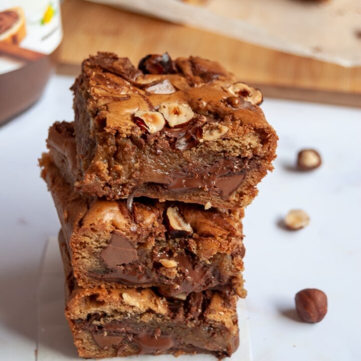 three blondie bars with Nutella and hazelnuts stacked on top of each other, a jar of nutella and more blondies on a wooden board