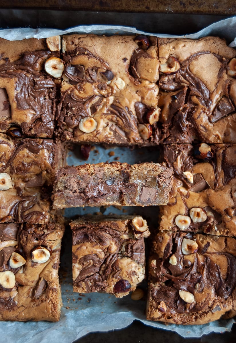 A close up picture of a tray of Nutella blondies topped with chocolate hazelnut spread and chopped hazelnuts.