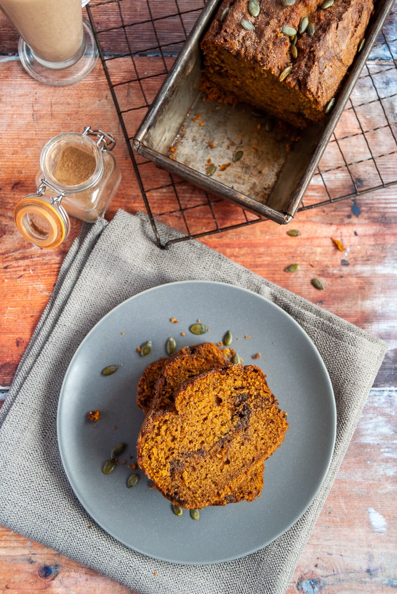 slices of pumpkin bread with a cinnamon swirl and sprinkled with pumpkin seeds on a grey/blue plate and grey napkin, pumpkin bread in a loaf tin on a wire rack and a little pot of pumpkin spice on a wooden backdrop