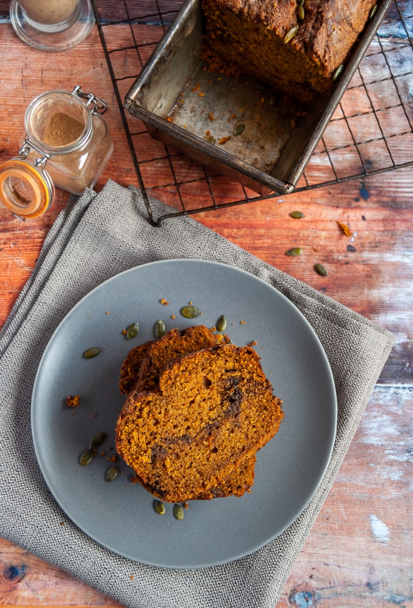 two slices of pumpkin loaf with a cinnamon swirl and pumpkin seeds on a grey plate on a grey napkin, pumpkin bread in a loaf tin on a wire rack and a small pot of pumpkin spice