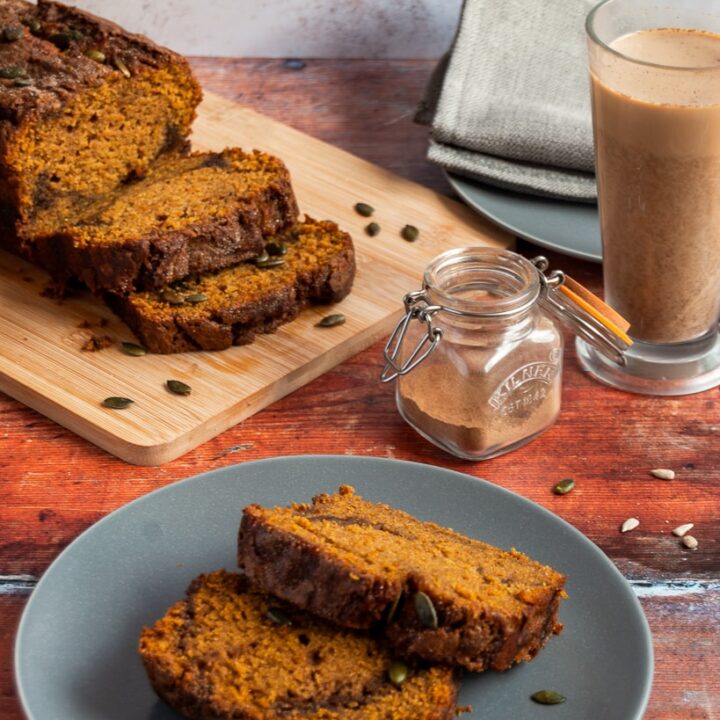 two slices of pumpkin loaf cake with a cinnamon swirl on a grey plate, a pumpkin loaf cake on a wooden board, a little pot of pumpkin spice and a cinnamon latte in a glass cup