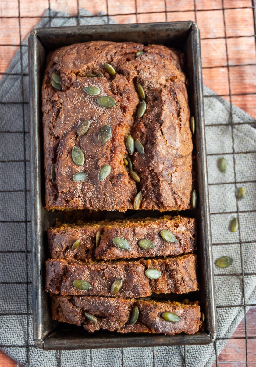 A partially sliced pumpkin loaf cake sprinkled with pumpkin seeds and cinnamon sugar in a loaf tin on a wire rack