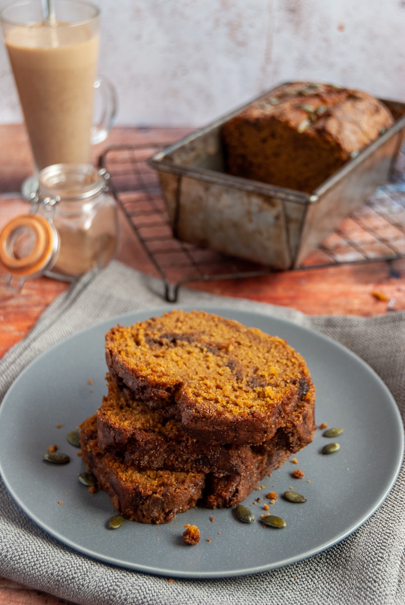 three slices of pumpkin loaf cake with a cinnamon swirl on a grey plate sprinkled with pumpkin seeds, a cinnamon latte in a glass cup and a pumpkin spice loaf on a wire rack