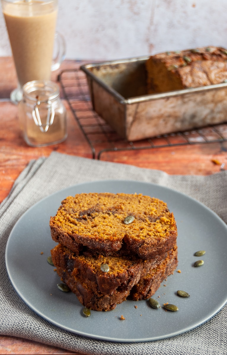 three slices of pumpkin bread on a grey plate, pumpkin bread in a loaf tin, a small pot of pumpkin spice and a cinnamon latte in a glass cup