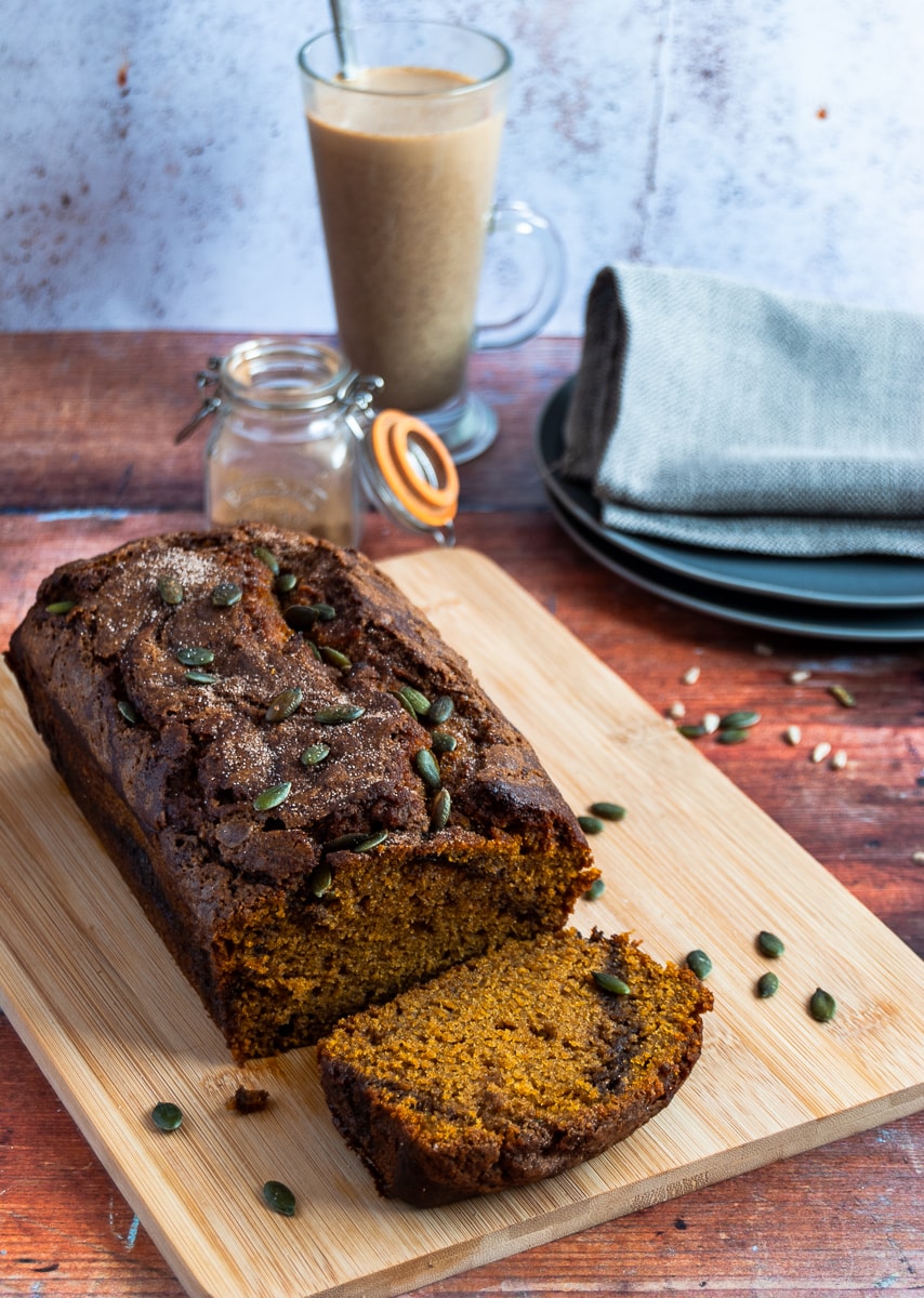 A pumpkin spice loaf cake sprinkled with pumpkin seeds on a wooden board, two grey plates with a linen napkin, a small pot of pumpkin spice and a cinnamon latte in a glass cup