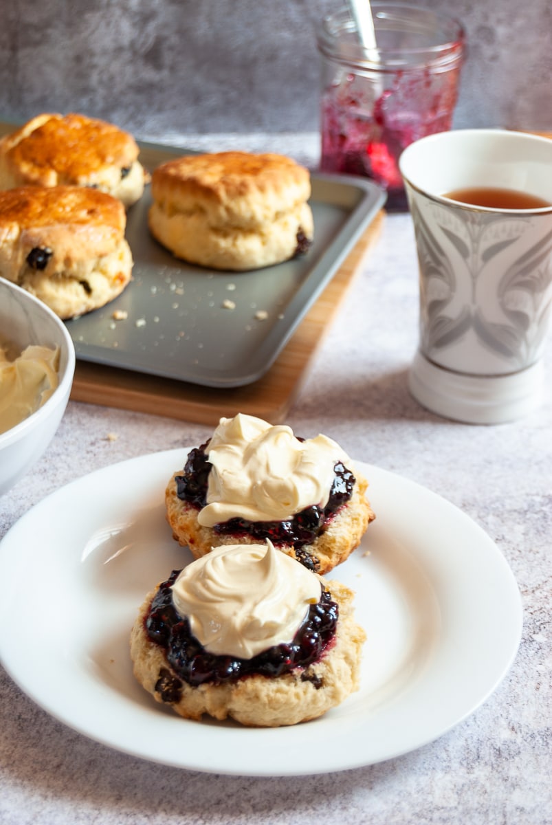 A white plate of fruit scones topped with whipped cream and fruit jam, a silver tray of scones, a white and silver cup of tea and a jar of jam with a spoon.