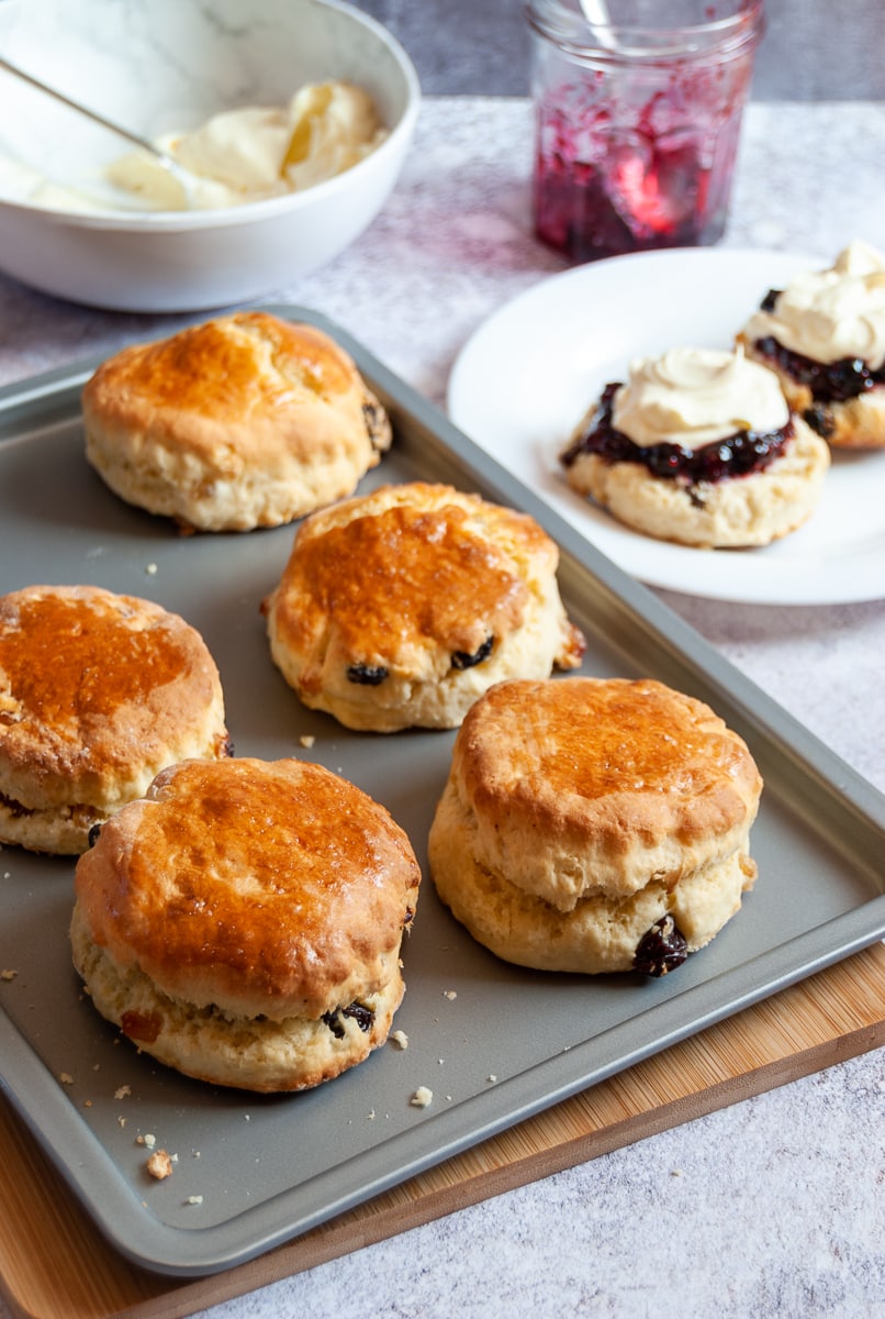 a silver baking tray of freshly baked fruit scones, a white plate with a scone cut in half topped with jam and cream, a jar of jam and a white bowl of whipped cream