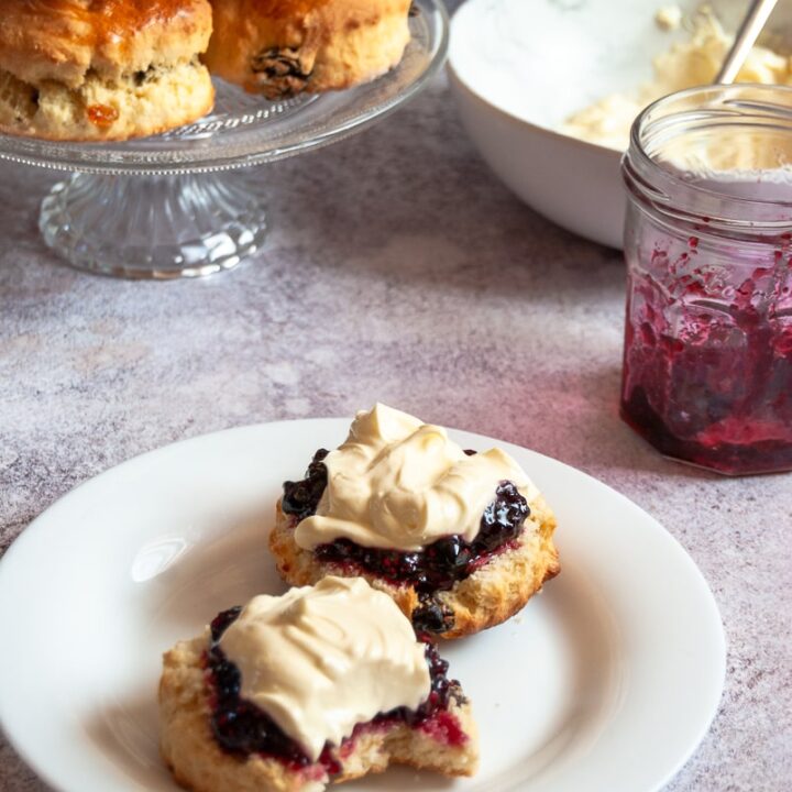 a white plate with a scone cut in half piled with whipped cream and mixed fruit jam, a jar of jam, a bowl of whipped cream and a glass serving plate with more scones piled on top