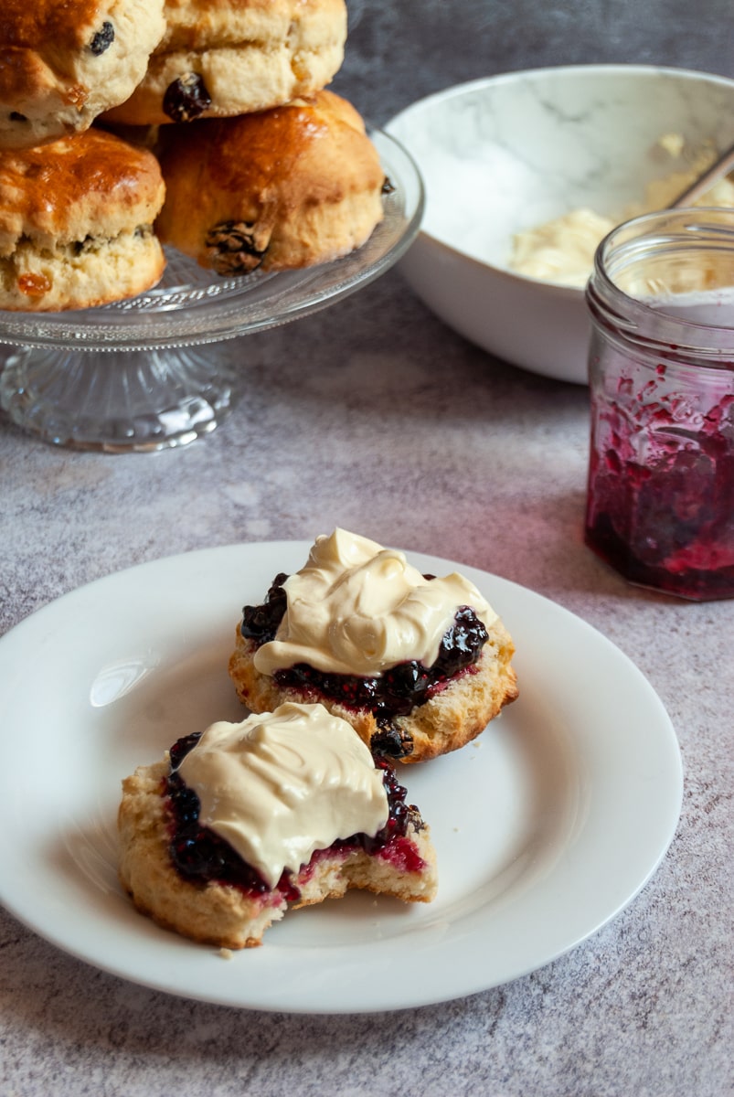 a white plate with a scone cut in half piled with whipped cream and mixed fruit jam, a jar of jam, a bowl of whipped cream and a glass serving plate with more scones piled on top