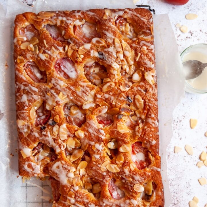 a flat lay photo of a plum and almond traybake cake on a piece of baking parchment, a jug of cream and three plums.