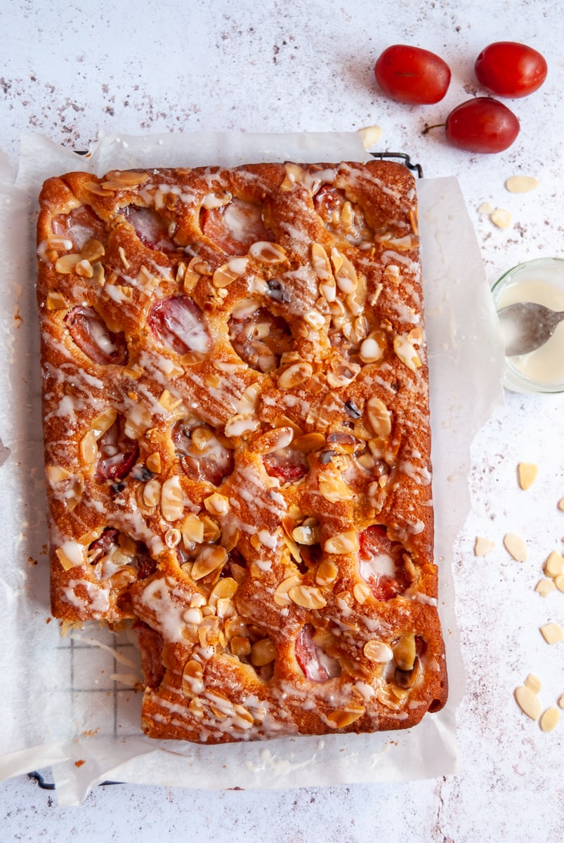 a flat lay photo of a plum and almond traybake cake on a piece of baking parchment, a jug of cream and three plums.