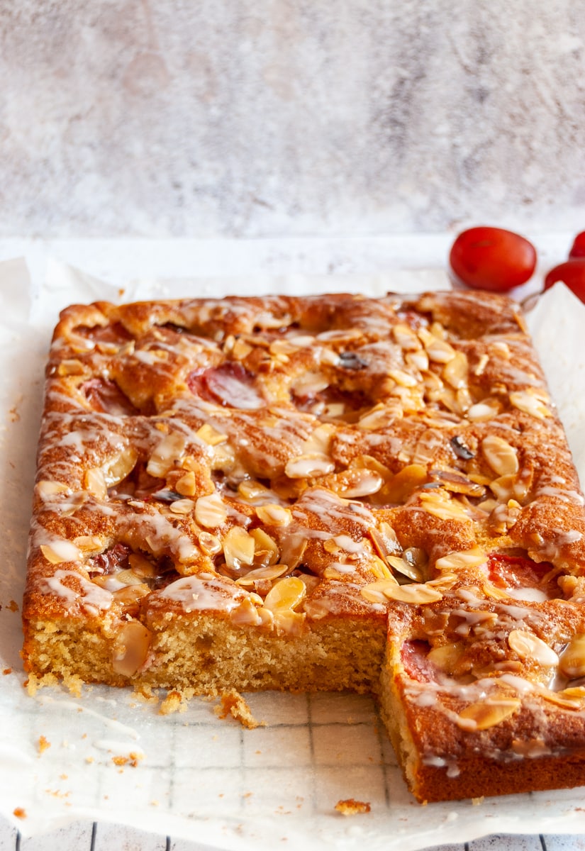 A plum and almond traybake topped with flaked almonds and an orange icing drizzle on a silver wire rack.