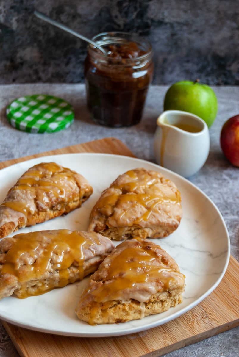 5 apple cinnamon scones drizzled with a caramel glaze on a white plate and wooden chopping board, a jar of apple butter with a spoon, a small jug of caramel sauce and a red and green apple.