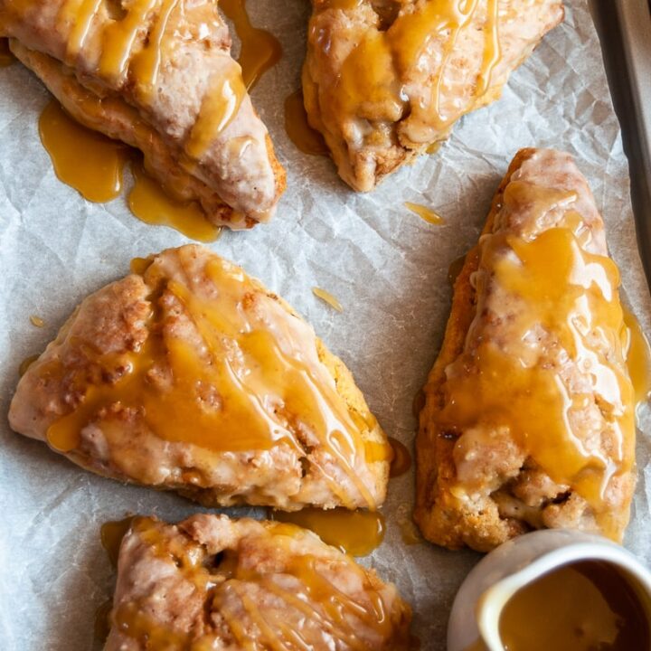 five apple and cinnamon scones drizzled with a caramel glaze on a baking tray and a small jug of caramel sauce